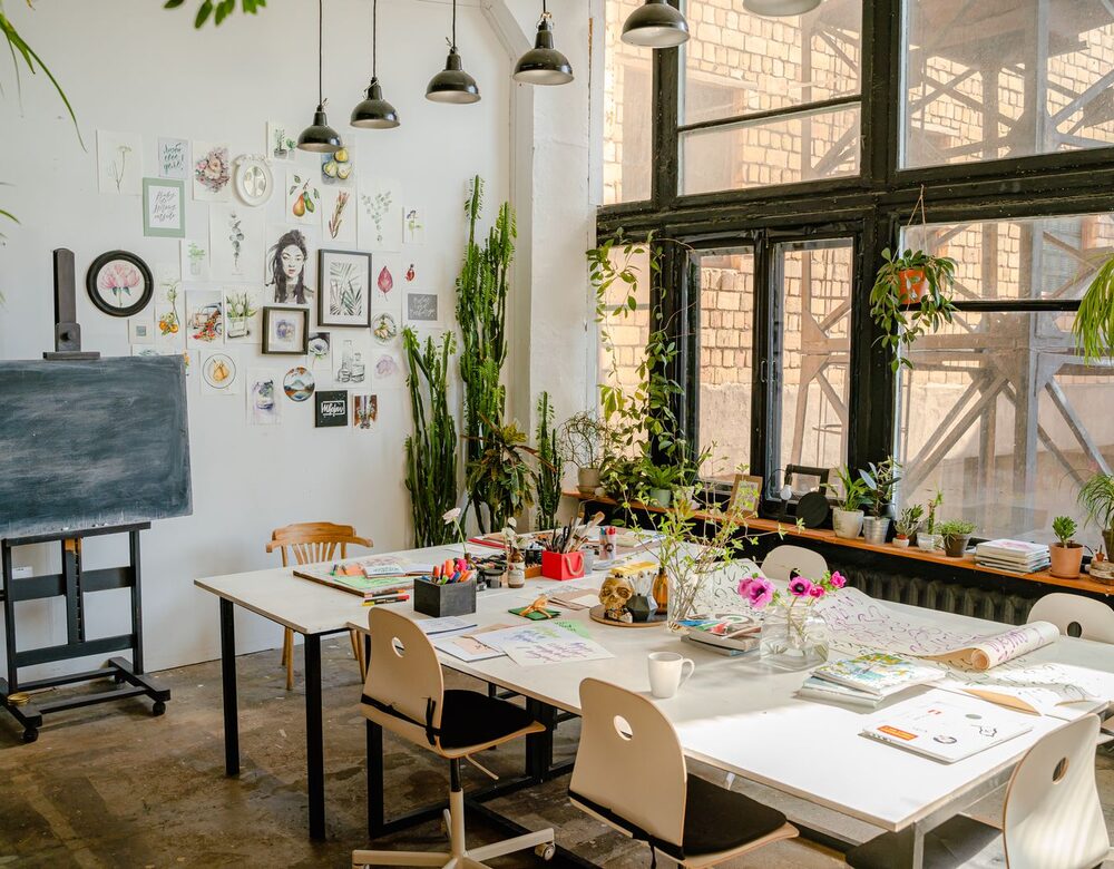 An airy creative studio with tall windows, plants, and a worktable of materials and sketches